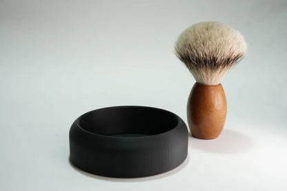Shaving brush with wooden handle and soft bristles next to a black shaving soap bowl for traditional wet shaving