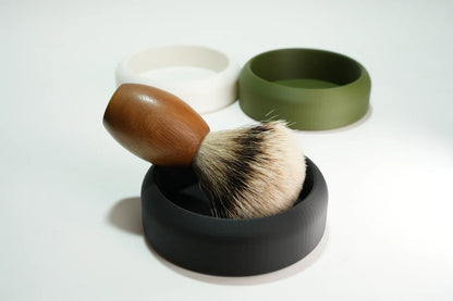 Shaving brush with wooden handle and soft bristles resting in a black silicone soap dish with white and green dishes in backg